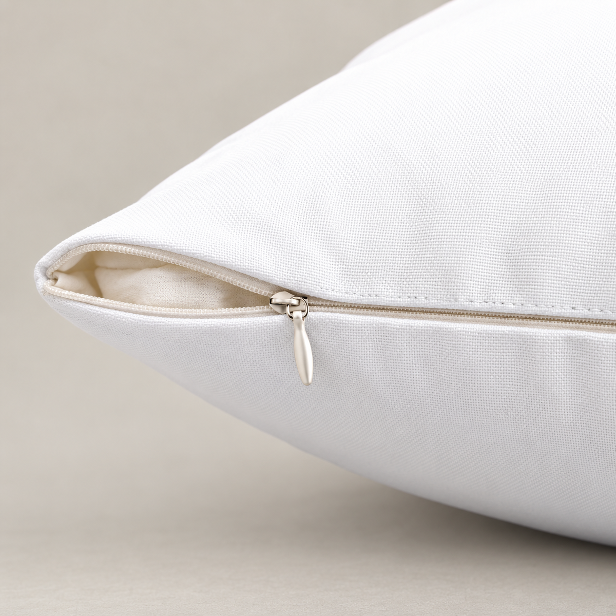 Close-up of a white pillow with a silver zipper, showing the fabric texture and interior lining against a neutral beige background.