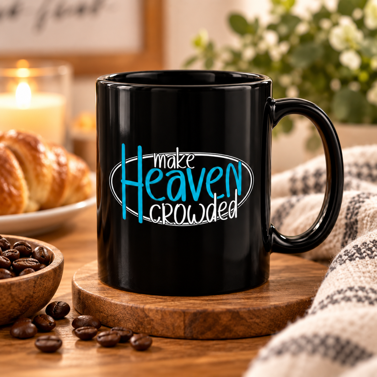 A black ceramic coffee mug with the text 'make Heaven crowded' in blue and white, sitting on a wooden coaster on a wooden table with coffee beans, a croissant, and a candle in the background.