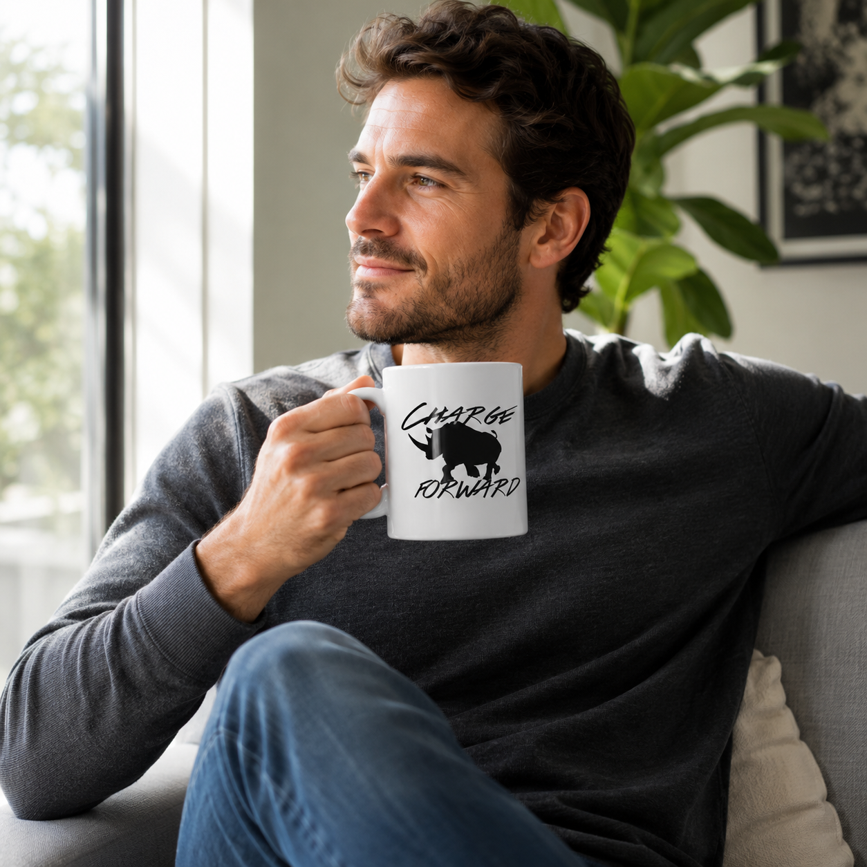 A man sitting on a couch in a bright living room, holding a white ceramic mug with a black rhinoceros silhouette and the words 'CHARGE FORWARD'.