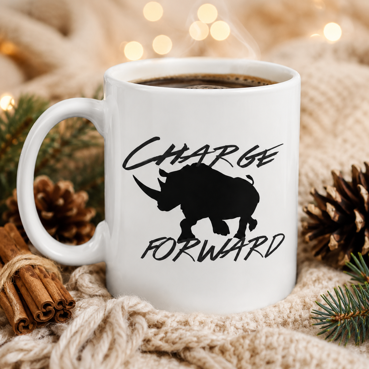 A white ceramic coffee mug with a black rhinoceros silhouette and the words 'CHARGE FORWARD' printed on the side, filled with coffee and styled on a knitted blanket with pinecones, cinnamon sticks, and warm string lights.