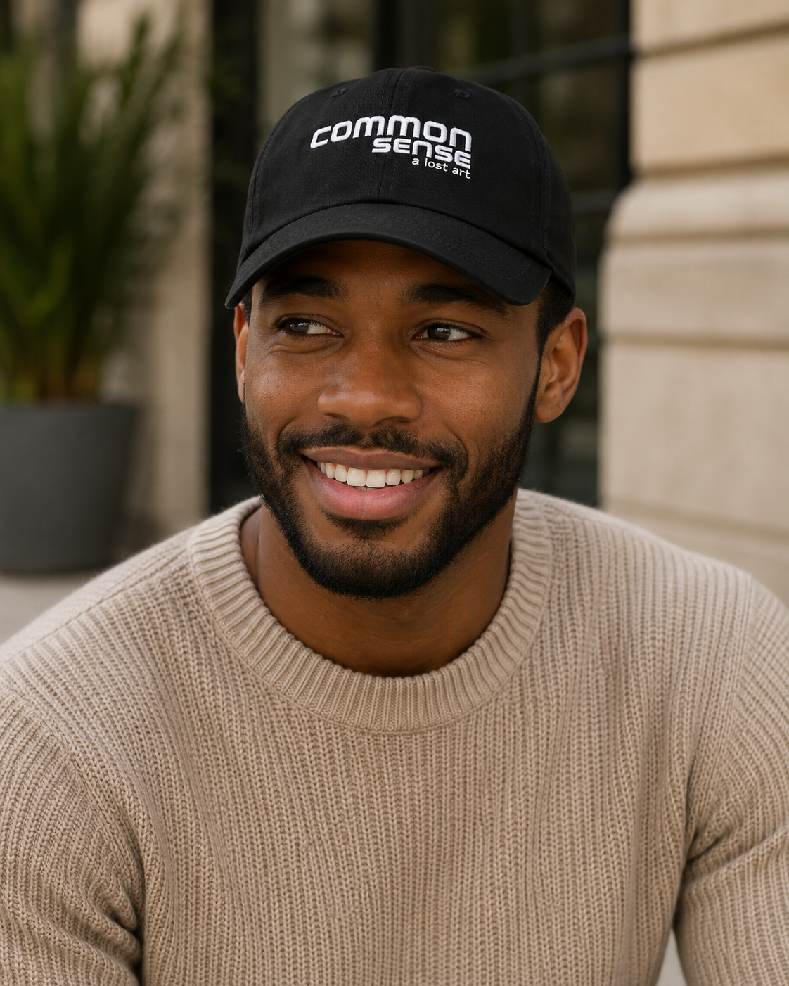 A smiling man wearing a black baseball cap with the white embroidered text 'common sense' and a beige knit sweater, photographed outdoors against a softly blurred building background.