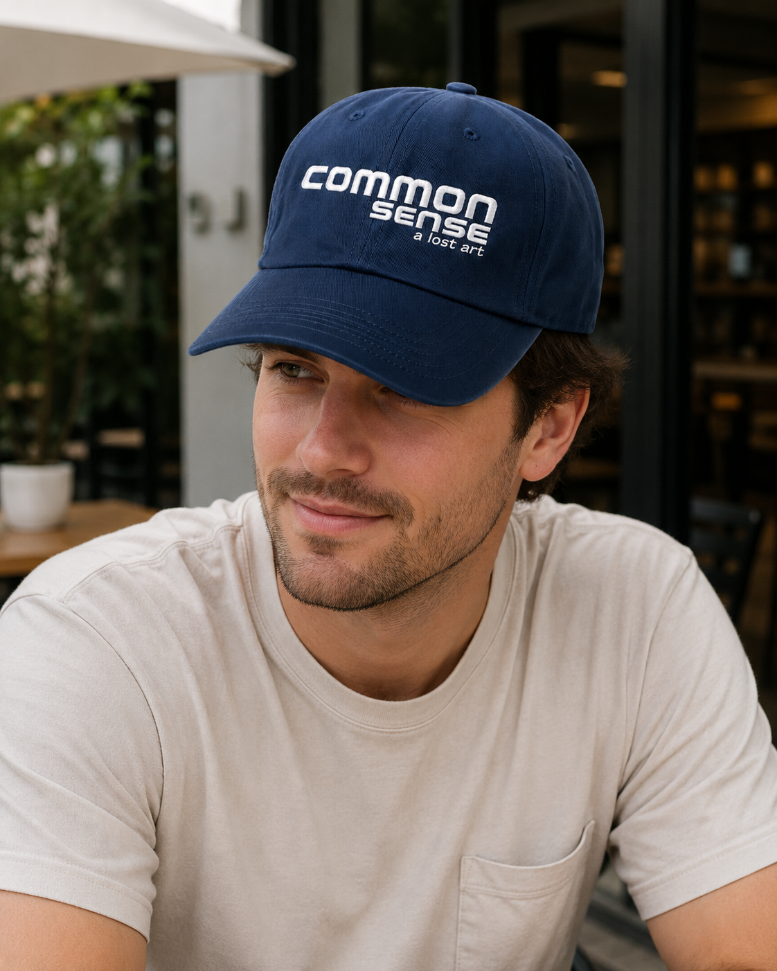 A man wearing a navy blue baseball cap with white embroidered text reading 'common sense' and 'A USE ART' in an outdoor cafe setting.