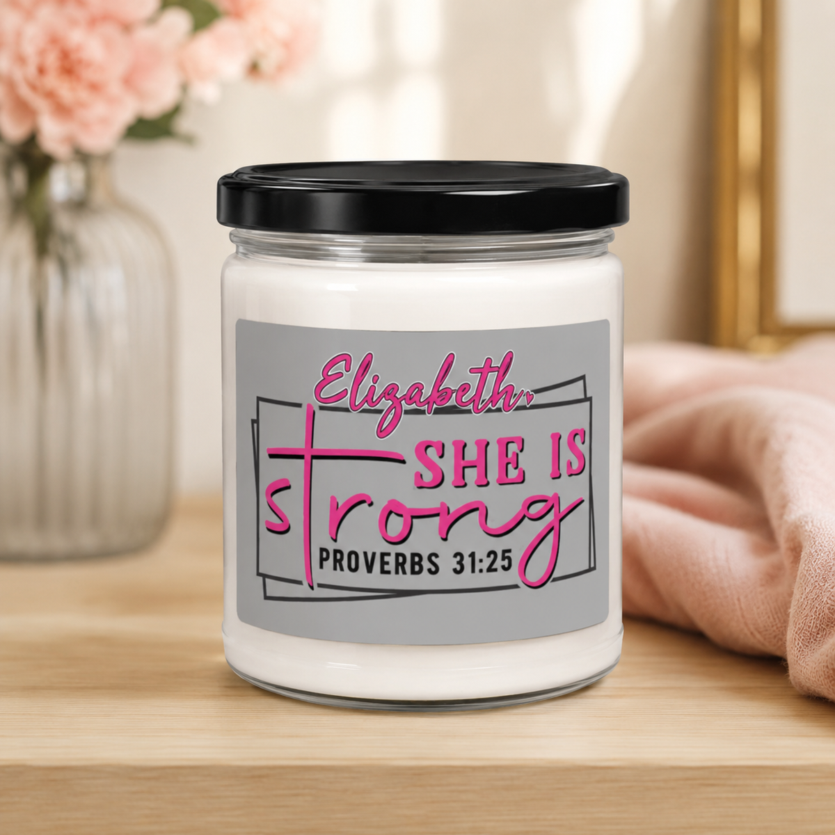 A white candle in a clear glass jar with a black lid and a gray label that reads 'Elizabeth, SHE IS strong PROVERBS 31:25' in pink and black text. The candle sits on a light wood surface with a blurred pink flower vase and soft pink fabric in the background.