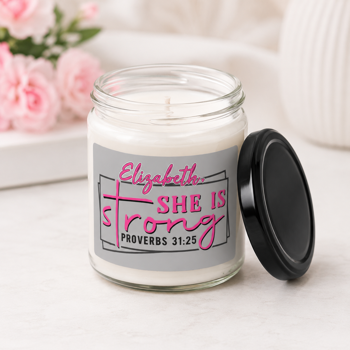 A personalized glass jar candle with a black lid resting beside it on a light marble surface. The candle has a gray label featuring the text 'Elizabeth SHE IS strong PROVERBS 31:25' in pink and black.