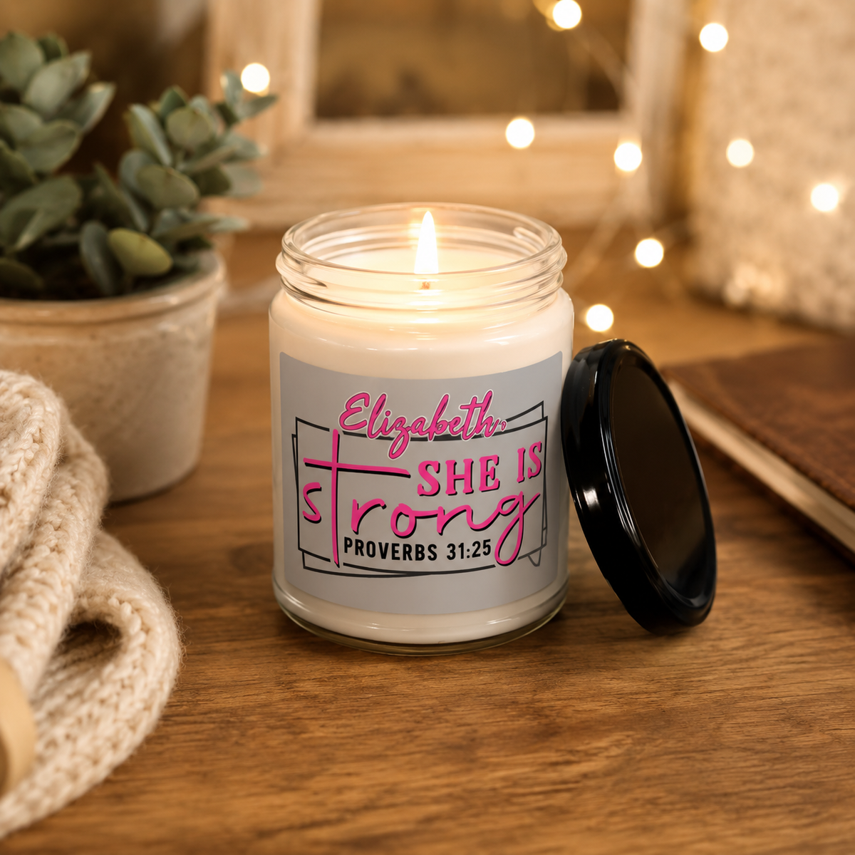 A lit white candle in a clear glass jar sits on a wooden table with its black lid beside it. The front label reads 'Elizabeth She Is Strong Proverbs 31:25' in pink and black text.
