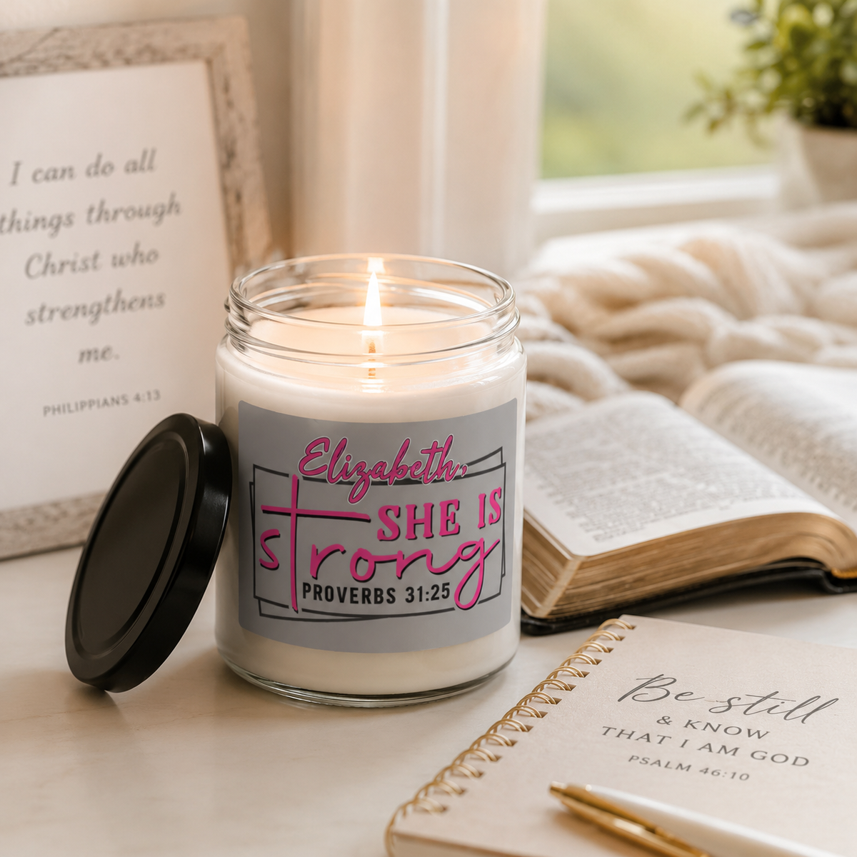 A personalized glass jar candle with a lit wick sits on a tabletop next to its black lid. The candle features a gray label with pink and black text reading 'Elizabeth She Is Strong Proverbs 31:25'.
