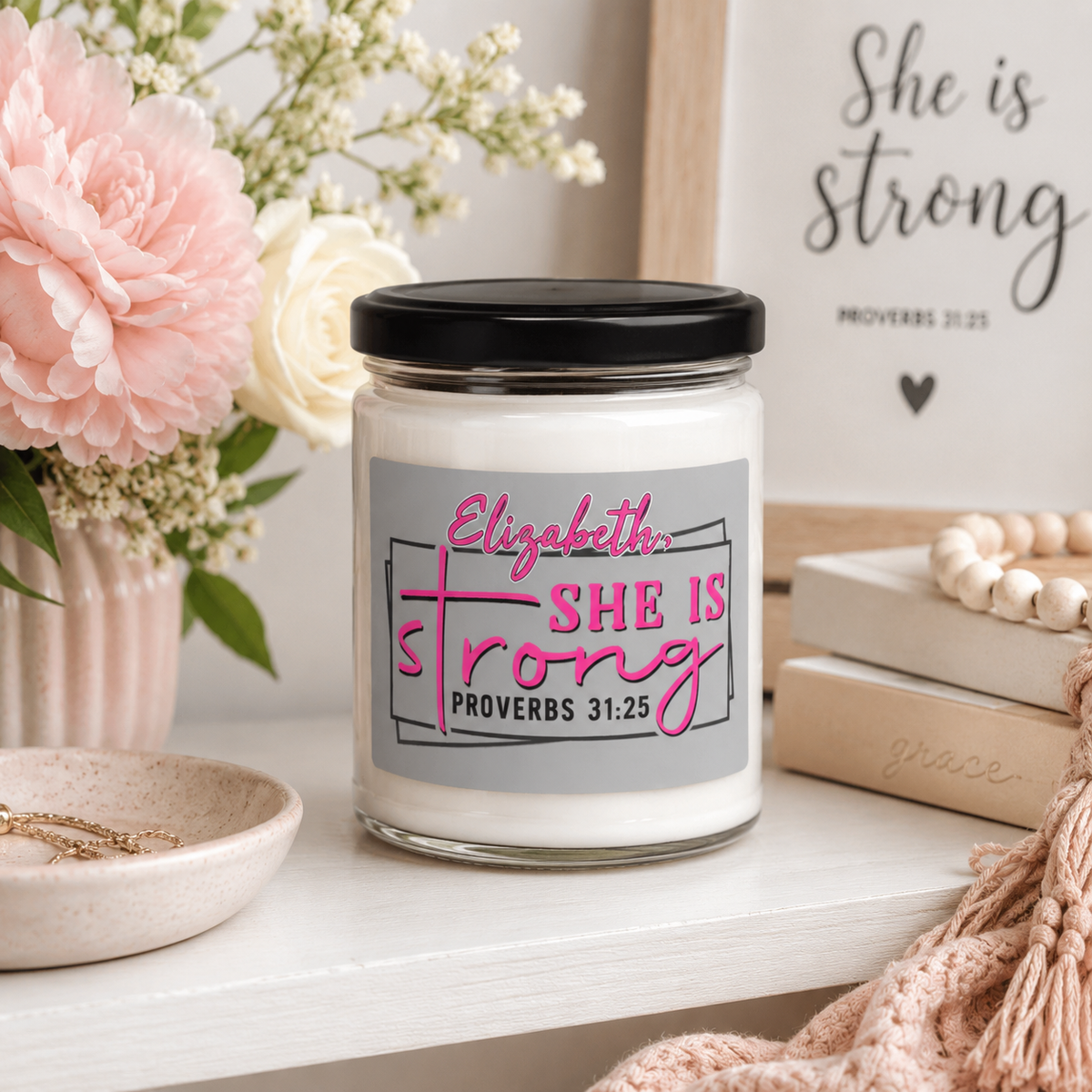 A personalized glass jar candle with a black lid and a gray label reading 'Elizabeth: SHE IS Strong PROVERBS 31:25' in pink and black lettering. The candle is styled on a white shelf with pink and white flowers, a wooden sign, books, and a knit blanket.