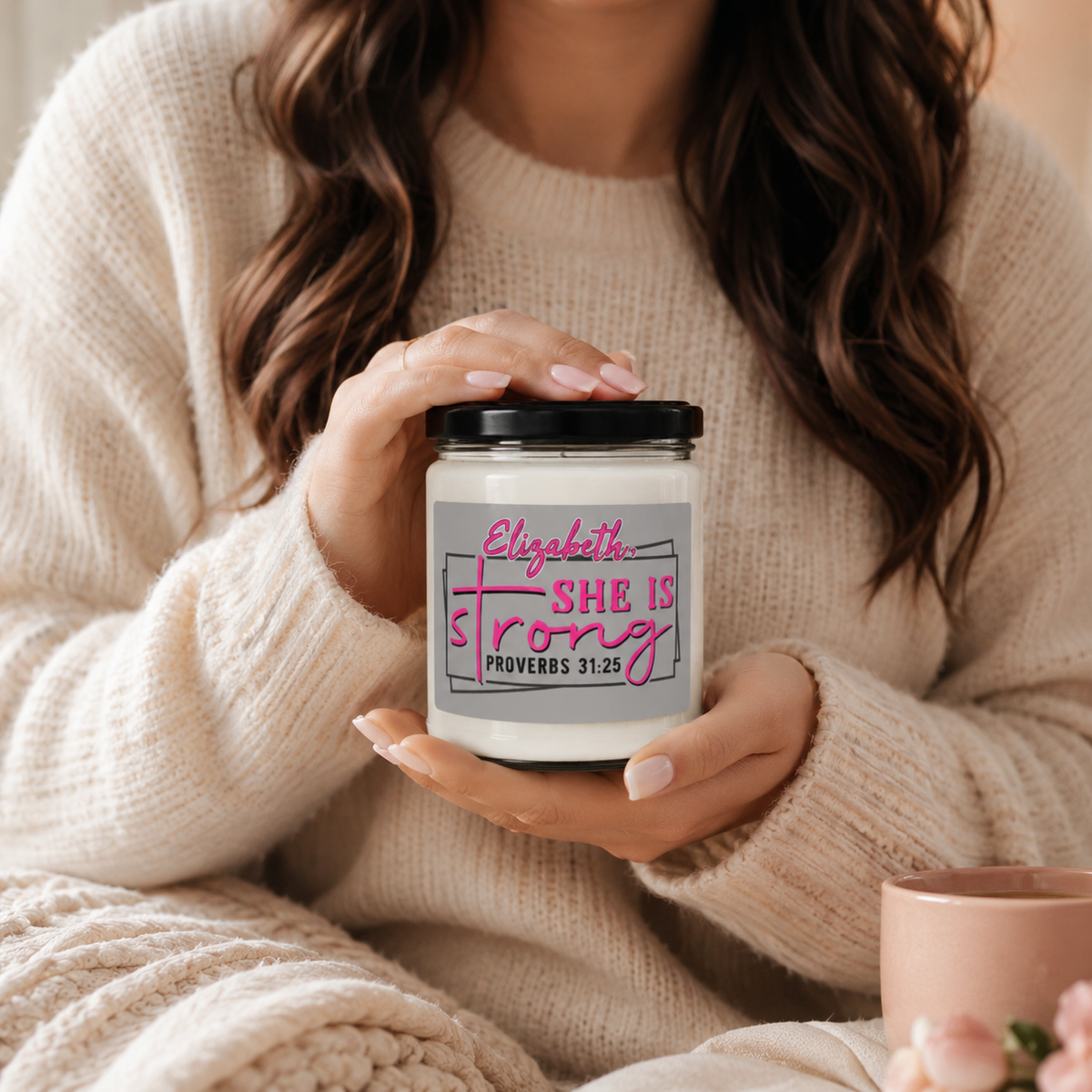 A woman in a cozy cream knit sweater holds a glass jar candle with a black lid and a gray label that reads 'Elizabeth She Is Strong Proverbs 31:25' in pink and black text.