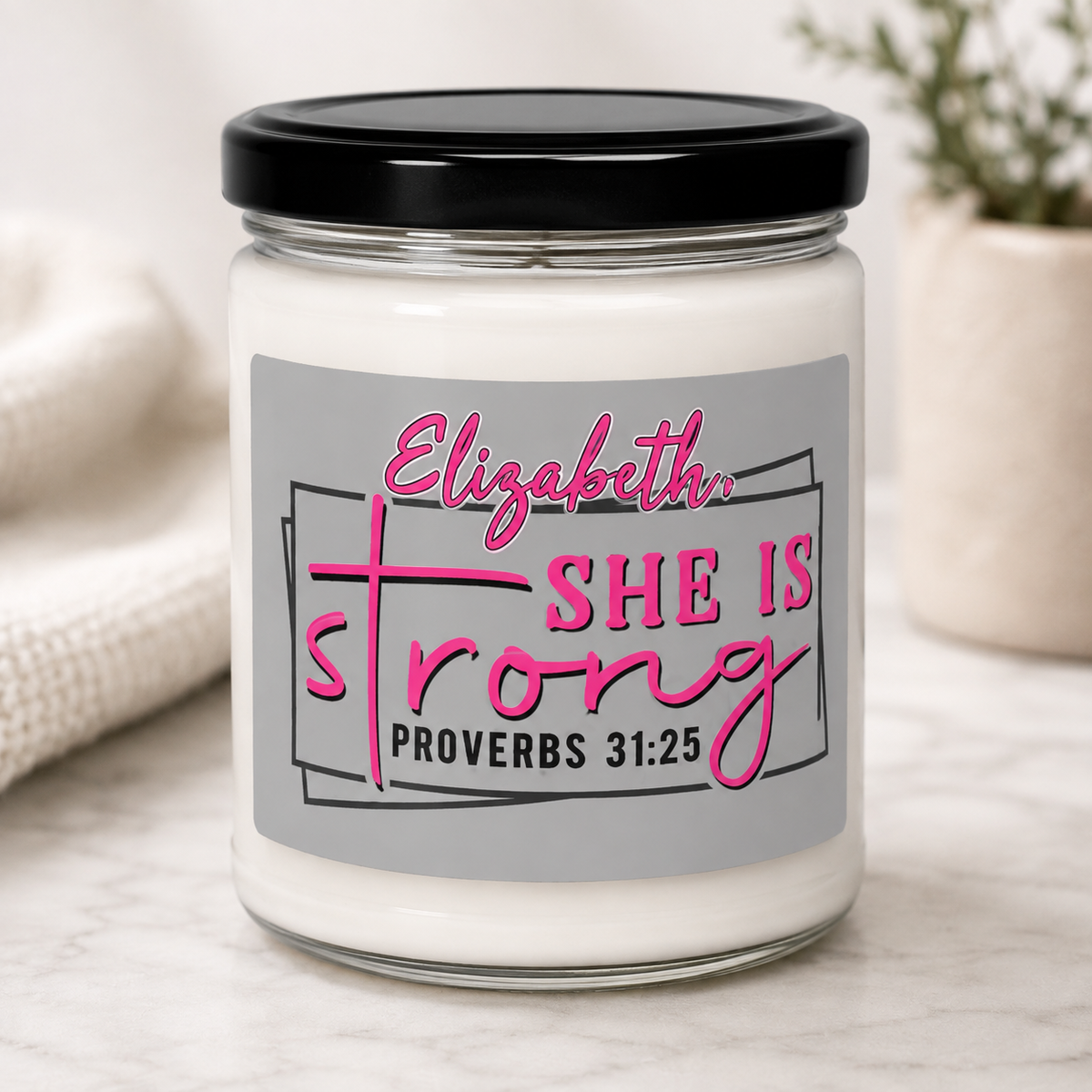 A personalized glass jar candle with a black lid and a gray label that reads 'Elizabeth, SHE IS strong PROVERBS 31:25' in pink and black lettering, set on a light marble surface with a soft neutral background.