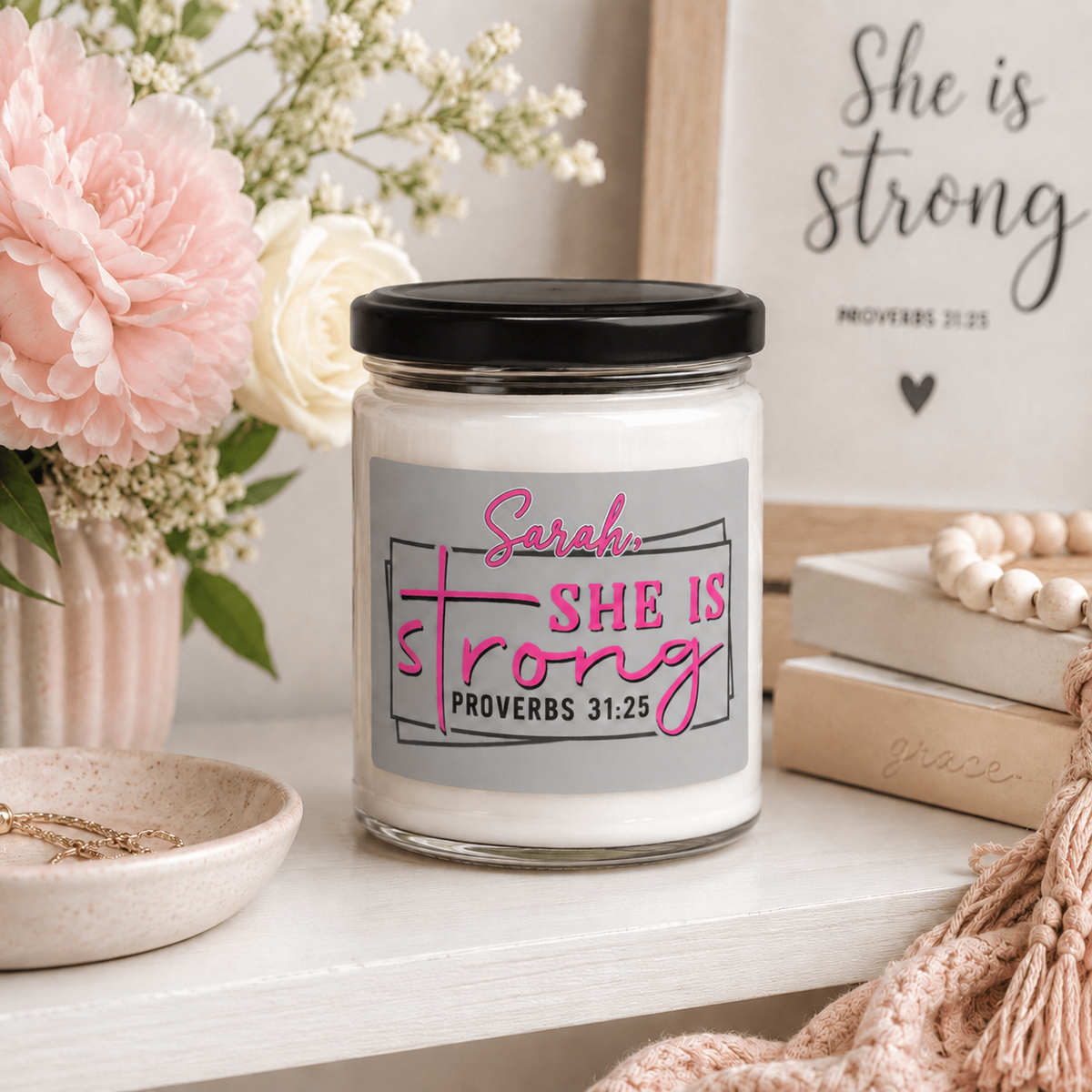 A personalized glass jar candle with a black lid and a gray label reading 'Sarah, SHE IS strong PROVERBS 31:25' in pink and black lettering. The candle is styled on a white shelf with pink and white flowers, a wooden sign, books, and a beige knit blanket.