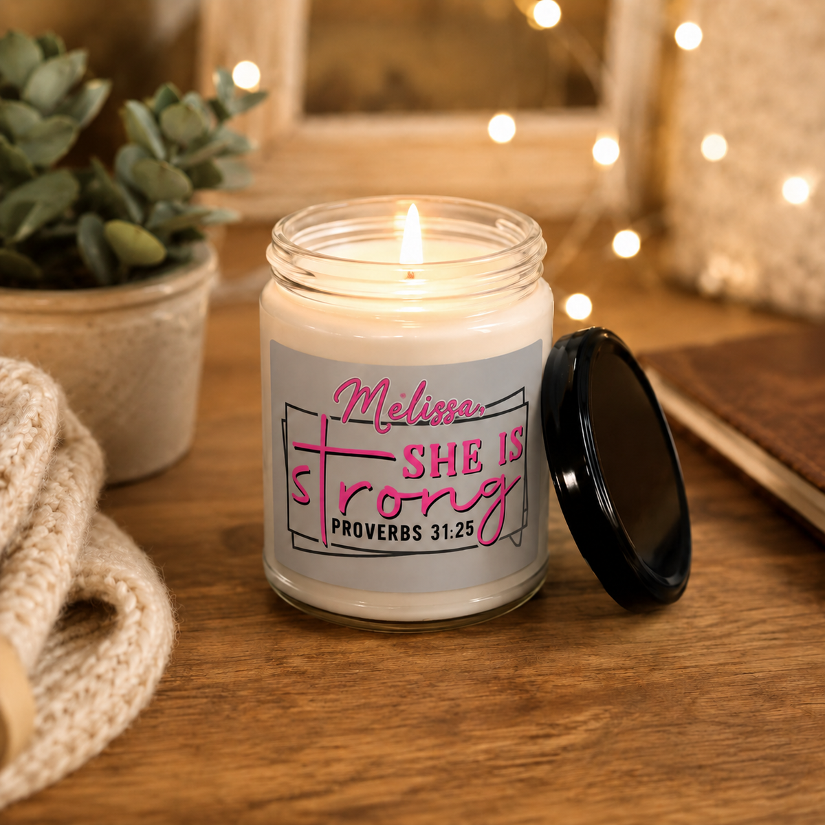 A lit white candle in a clear glass jar with a black lid resting beside it on a wooden table. The label reads 'Melina SHE IS Strong PROVERBS 31:25' in pink and black text.