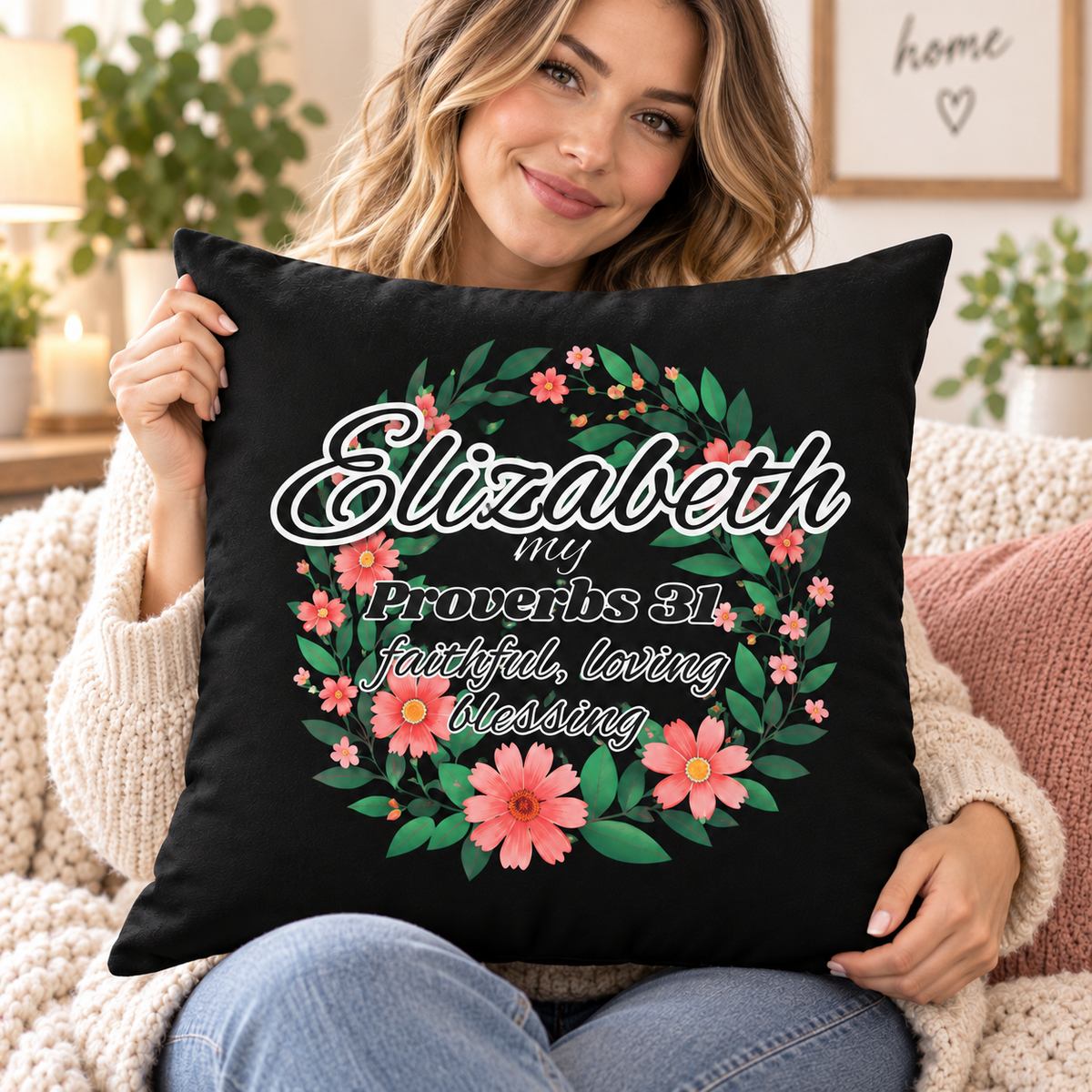 A woman sitting on a cozy knit couch holds a black square throw pillow featuring a floral wreath design and the text 'Elizabeth my Proverbs 31:10 faithful, loving blessing' in white script and serif fonts.