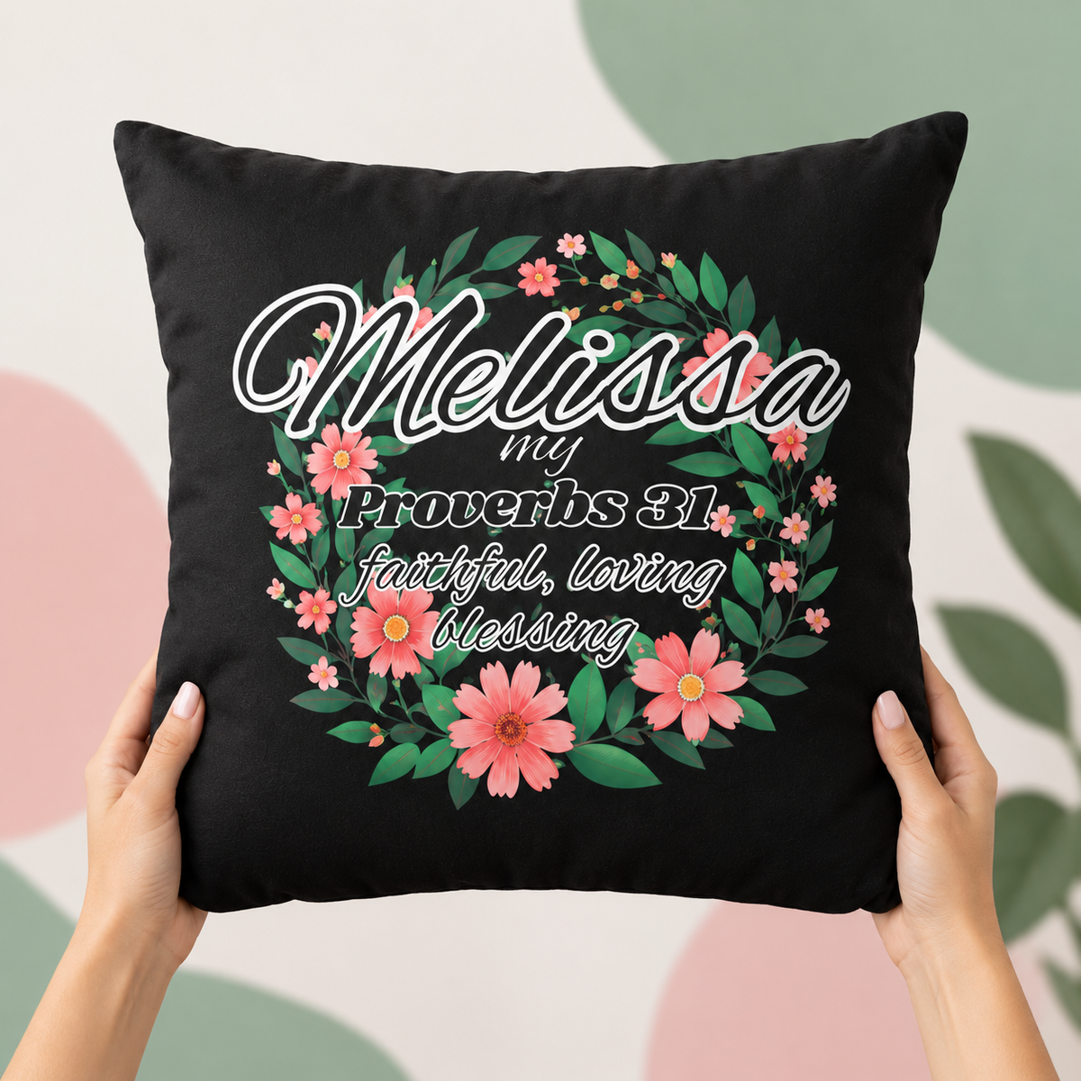 A black square throw pillow held by two hands, featuring a floral wreath design with pink flowers and green leaves, and the text 'Melissa my Proverbs 31 faithful, loving Blessing'. The background is a soft, abstract pattern of pastel pink, green, and white.
