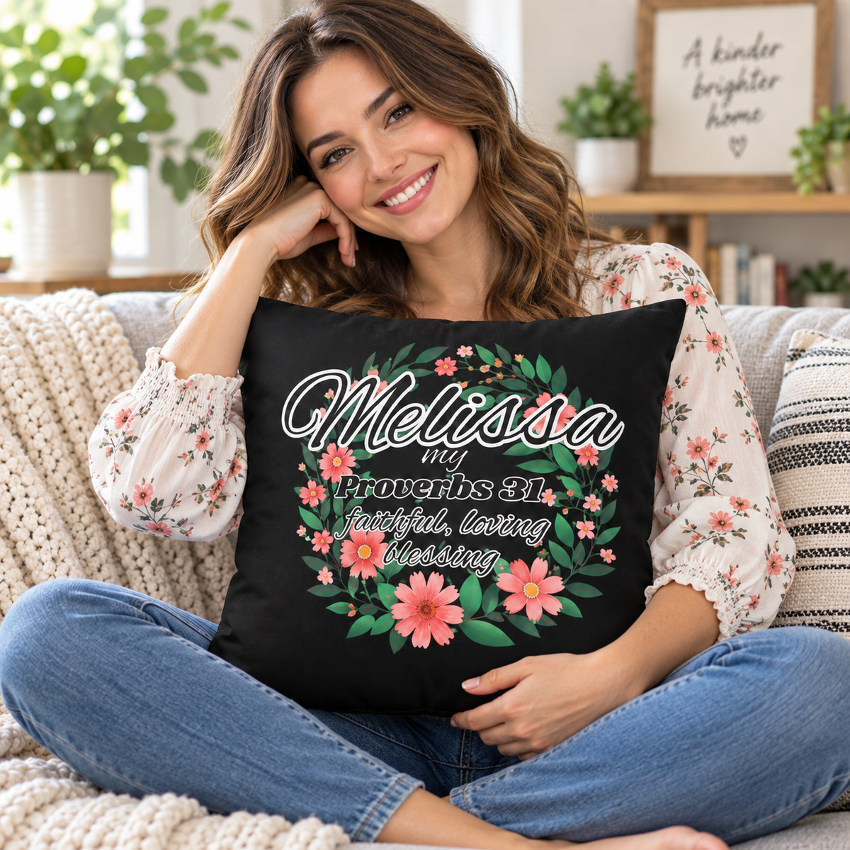 A woman sitting on a cozy couch holds a black square throw pillow featuring a personalized floral wreath design with the text 'Melissa is faithful, loving goddess' from Proverbs 31:25. The scene is set in a bright, modern living room with plants and home decor in the background.