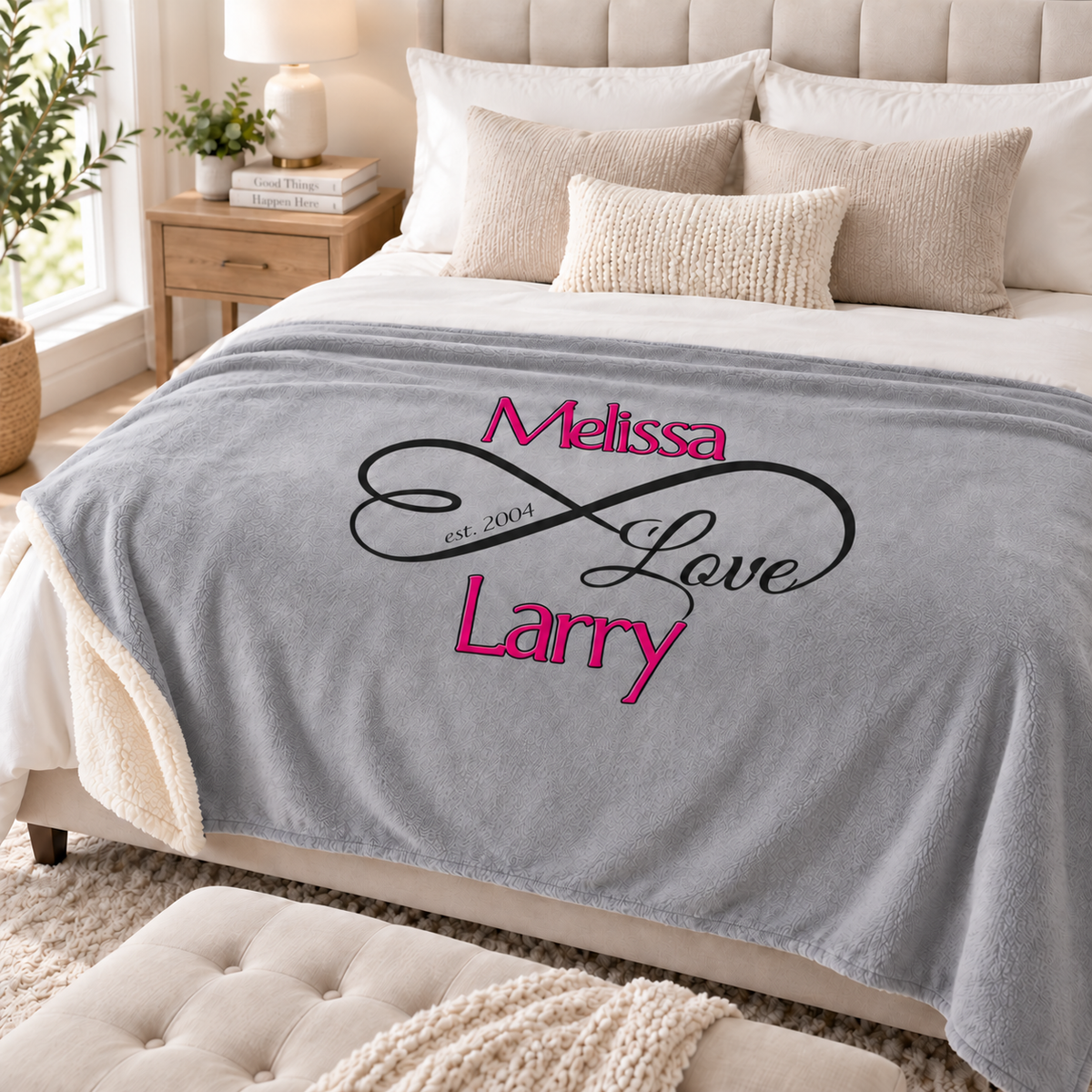 A personalized gray fleece throw blanket draped over a bed in a bright, modern bedroom. The blanket features a black infinity symbol with the words 'Melissa Love Larry' in pink script and 'est.