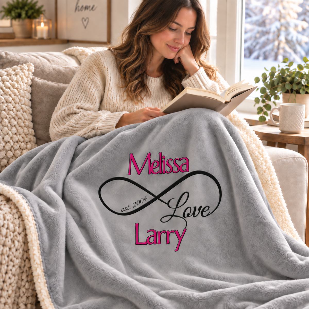 A woman in a cream knit sweater sits on a couch reading a book, wrapped in a gray plush blanket printed with the words 'Melissa Love Larry' in pink and black script.
