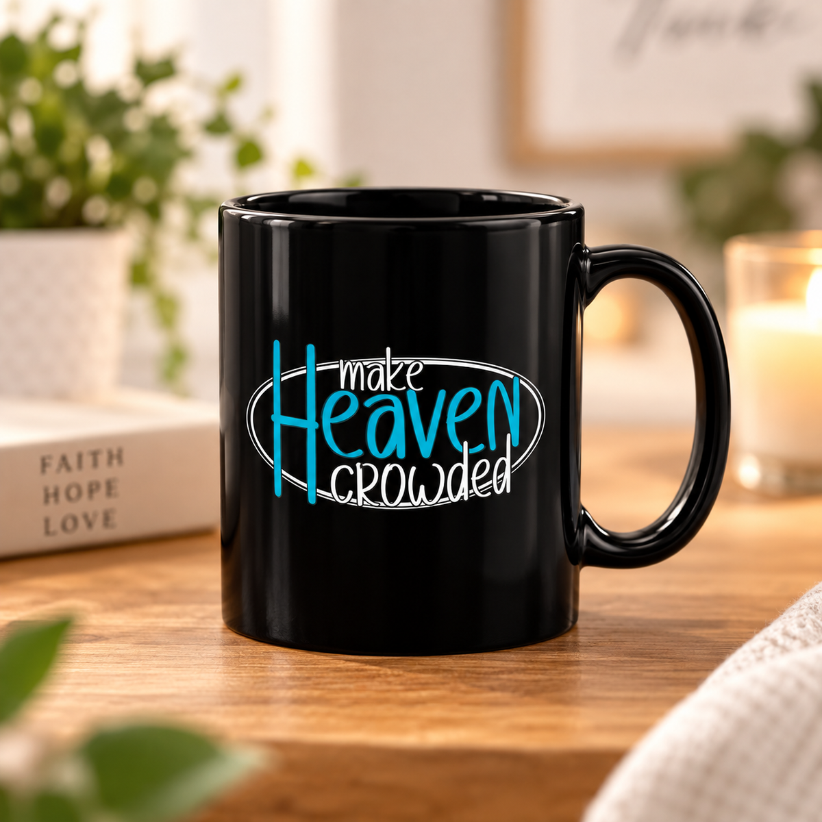 A black ceramic mug with the printed phrase 'make Heaven crowded' in white and bright blue lettering, sitting on a wooden table in a cozy indoor setting with a blurred candle, book, and plant in the background.