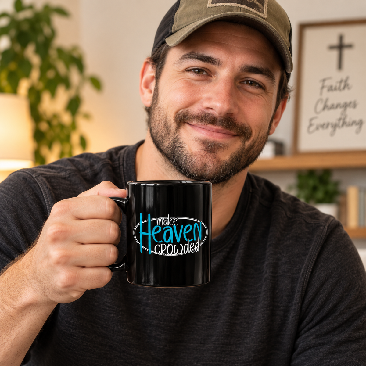 A smiling man in a dark gray t-shirt and baseball cap holds up a black ceramic mug with the text 'make Heaven crowded' in white and bright blue script. The background shows a warm, cozy home interior with a framed sign that reads 'Faith Changes Everything'.
