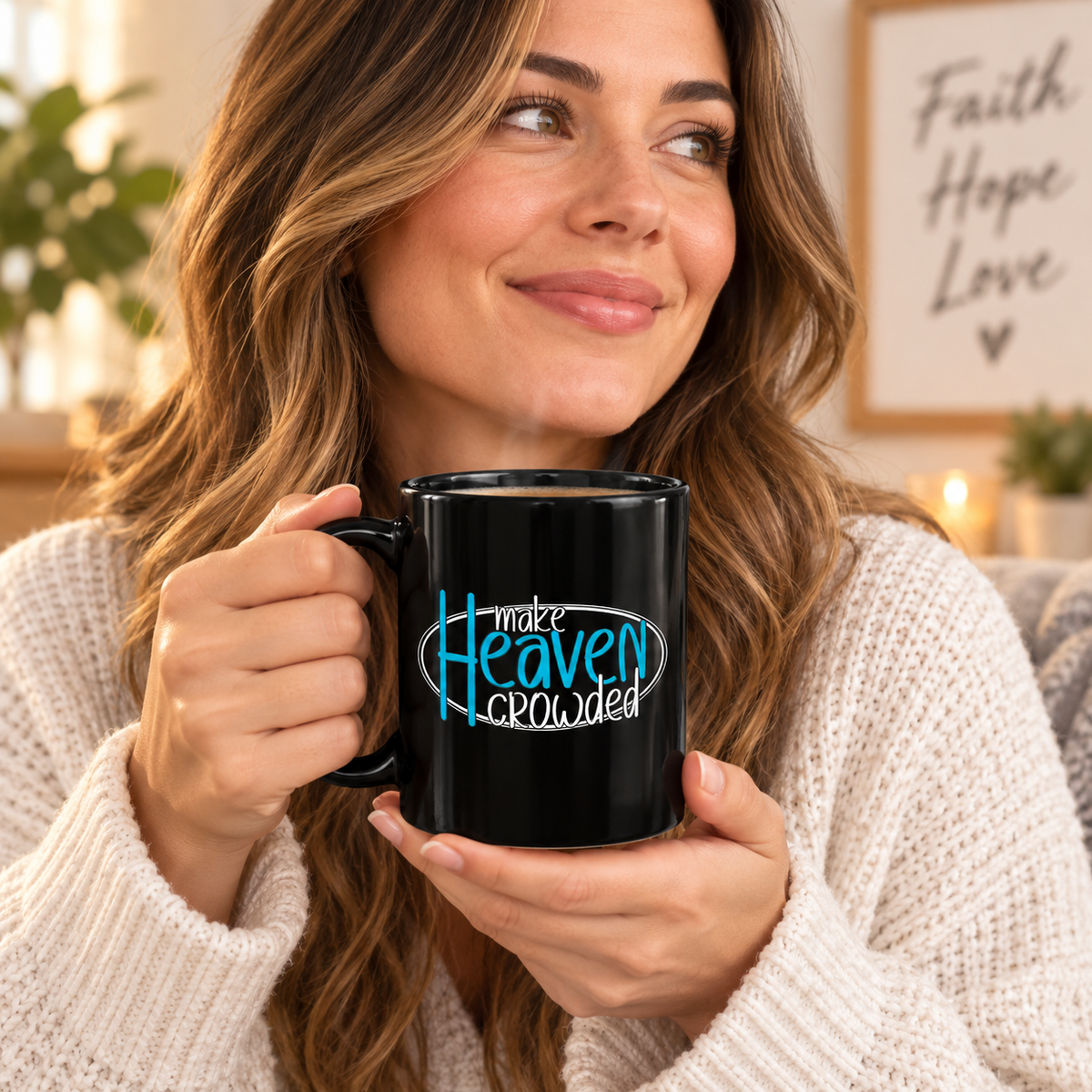 A woman in a cream knit sweater holds a black ceramic mug with the text 'make Heaven crowded' in blue and white. The background shows a cozy, warm indoor setting with a framed sign that reads 'Faith Hope Love'.