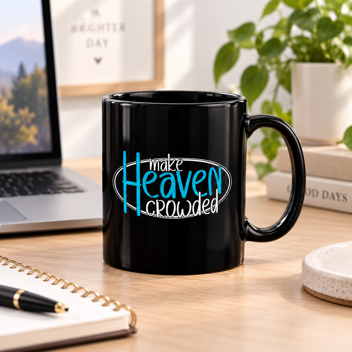 A black ceramic coffee mug with the text 'make Heaven crowded' in blue and white, sitting on a wooden desk in a styled home office setting with a laptop, notebook, and plant in the background.