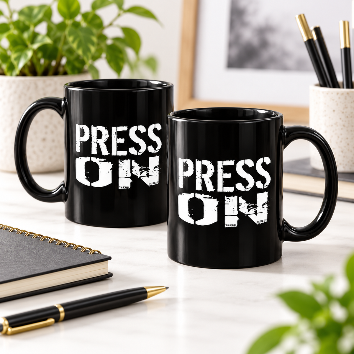 Two black ceramic mugs with the phrase 'PRESS ON' printed in distressed white letters, displayed on a light marble desk with office props including a notebook, pen, and potted plant.