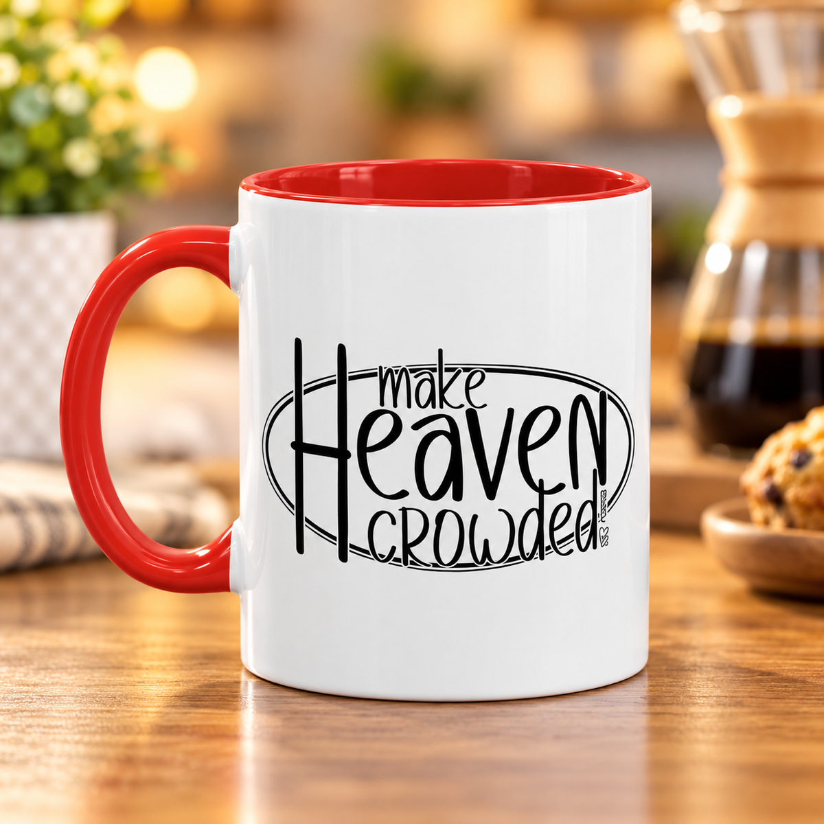 A white ceramic coffee mug with a red handle and red interior, featuring the black printed text 'make Heaven crowded' on the front, set on a wooden table with a blurred coffee maker in the background.