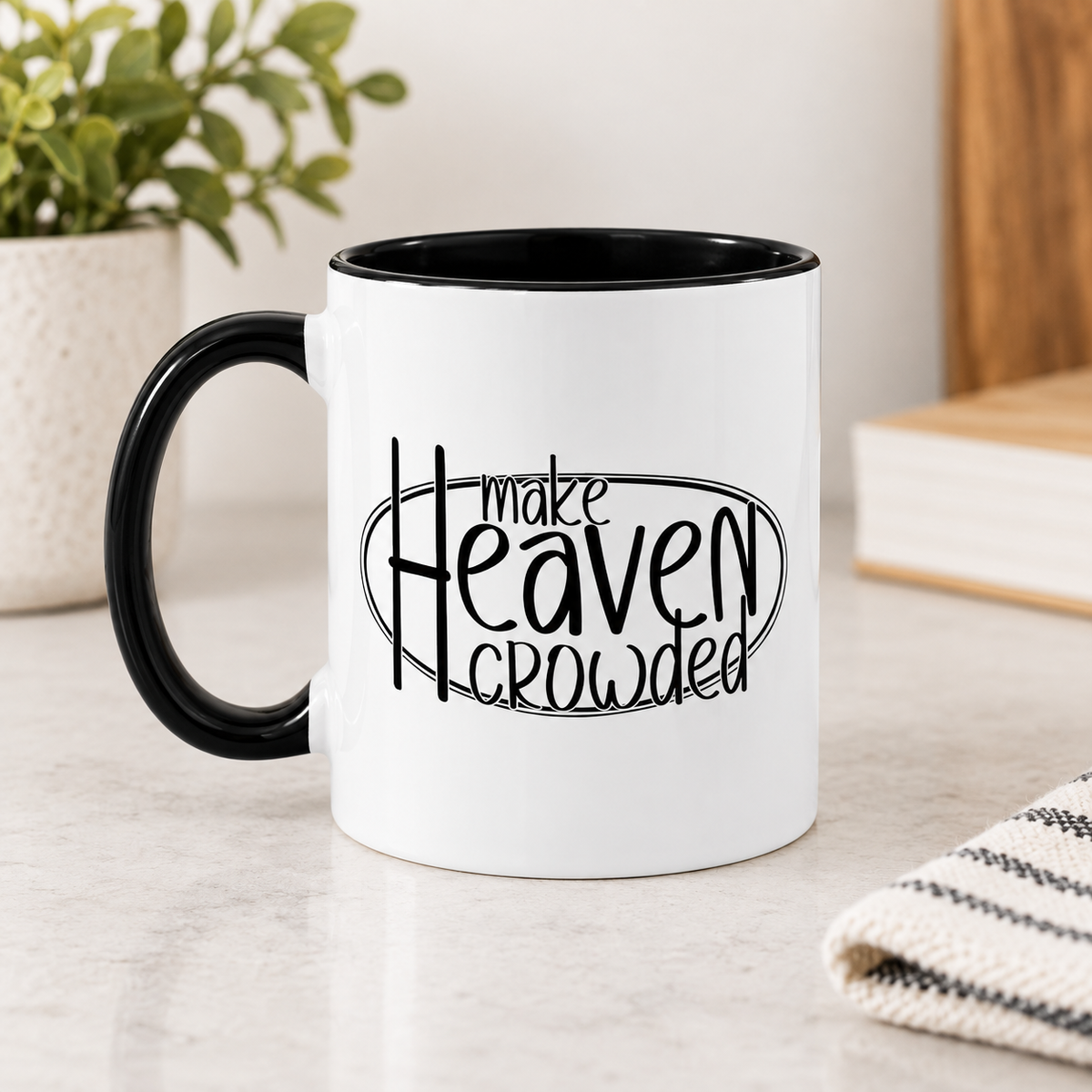 A white ceramic coffee mug with a black handle and black interior, featuring the printed phrase 'make Heaven crowded' in black lettering. The mug is set on a light marble countertop with a blurred potted plant and books in the background.