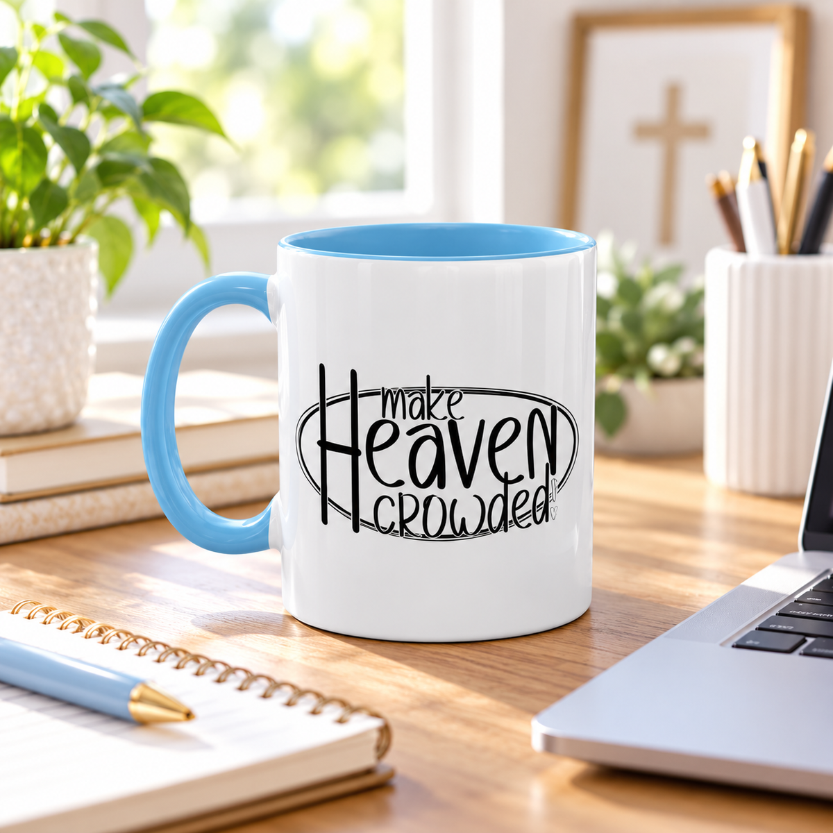 A white ceramic coffee mug with a light blue handle and interior, printed with the phrase 'make Heaven crowded' in black script and block lettering, sitting on a wooden desk in a bright, modern room.