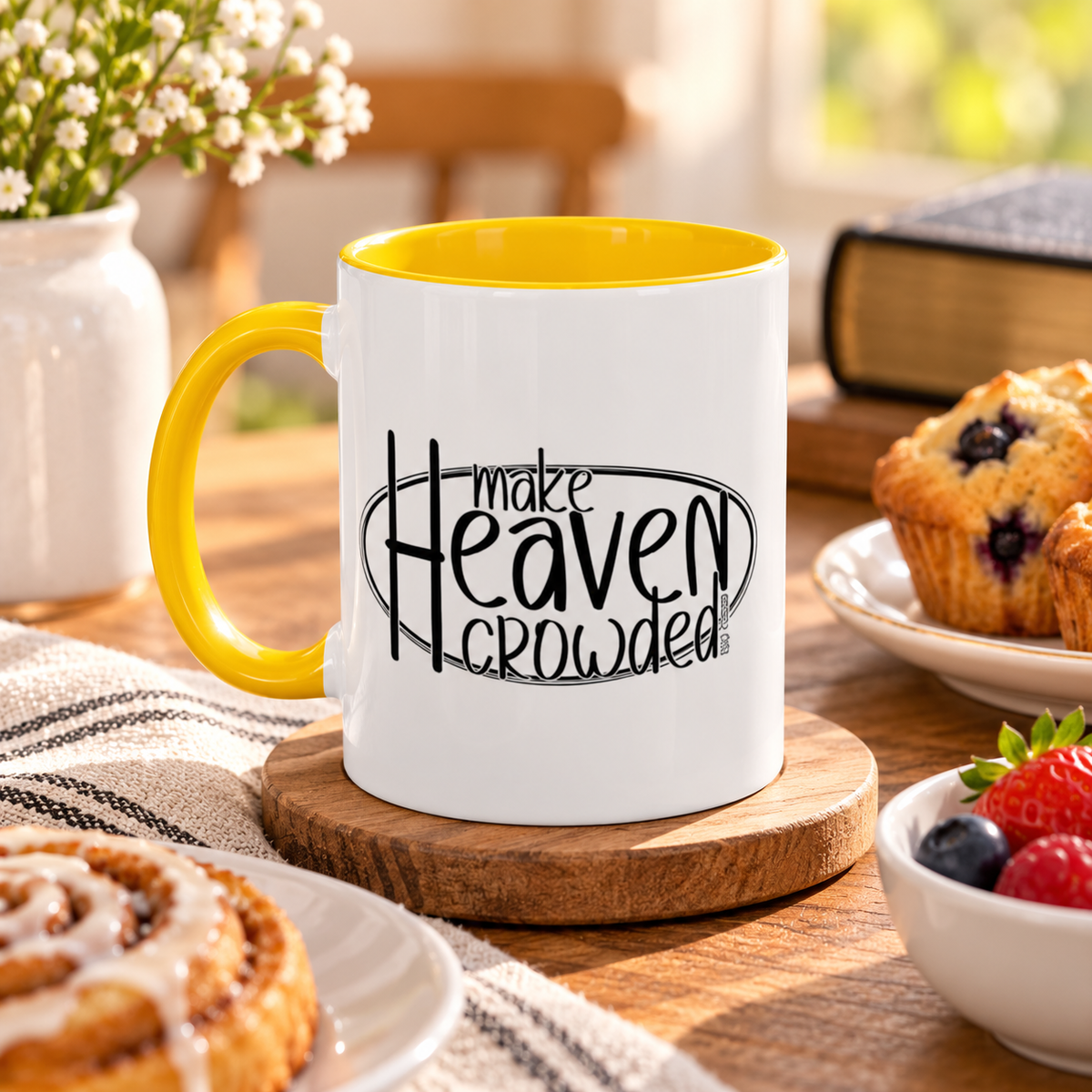 A white ceramic coffee mug with a yellow handle and interior, featuring the black printed text 'make Heaven crowded' on a wooden coaster on a breakfast table surrounded by muffins, berries, and a cinnamon roll.