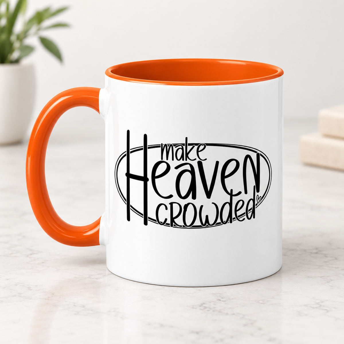 A white ceramic coffee mug with a bright orange handle and interior, featuring the black printed text 'make Heaven crowded' on the front, set on a light marble surface.