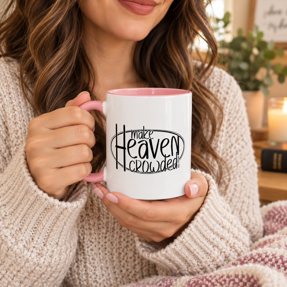 A woman in a beige knit sweater holds a white ceramic mug with a pink interior and handle. The mug has a black graphic that reads 'make Heaven crowded' in stylized lettering.