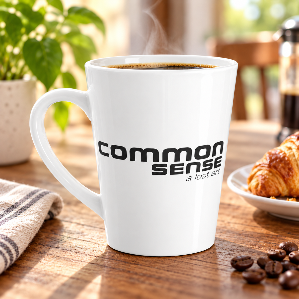 A white ceramic coffee mug with the words 'common sense a lost art' printed in black, filled with black coffee and set on a wooden table with a croissant, coffee beans, and a plant in the background.