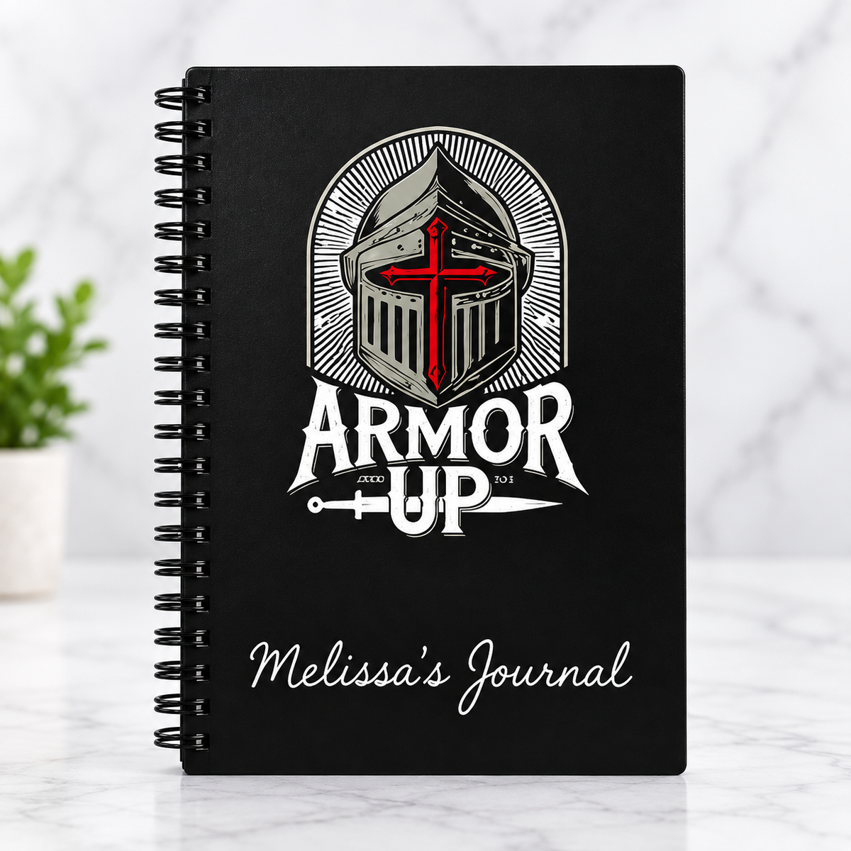 A black spiral-bound journal standing upright on a light marble surface. The cover features a white knight helmet graphic with a red cross, the words 'ARMOR UP', and 'Melissa's Journal' in white script.