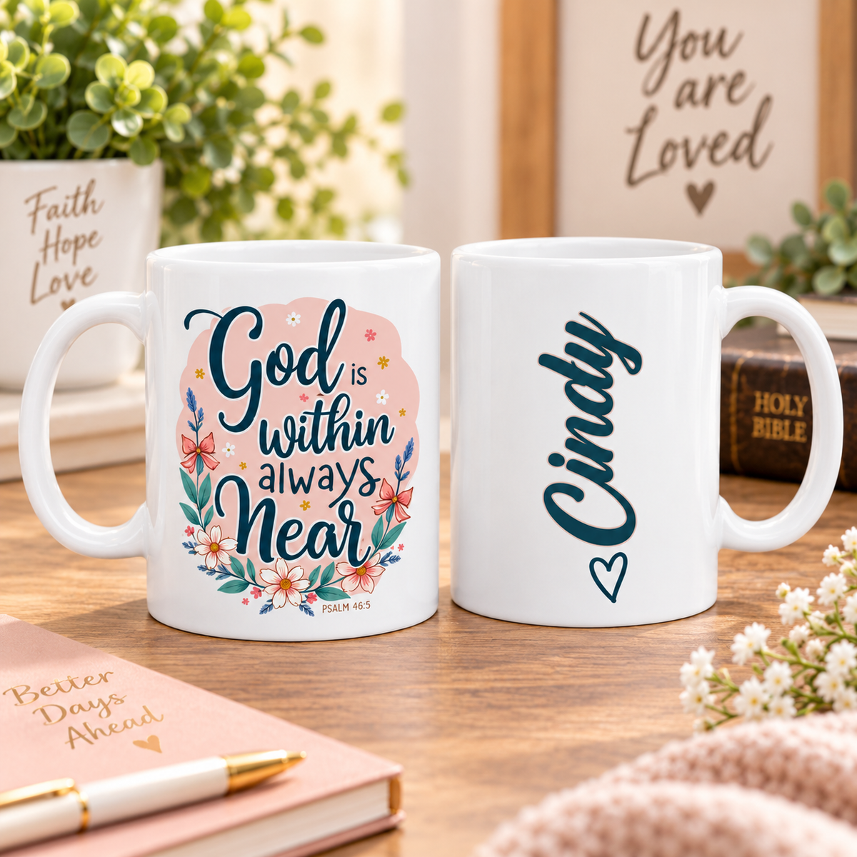 Two white ceramic mugs displayed on a wooden table in a styled home setting. The left mug features a pink and teal floral graphic with the text 'God is within always near' and 'Psalm 46:5'.