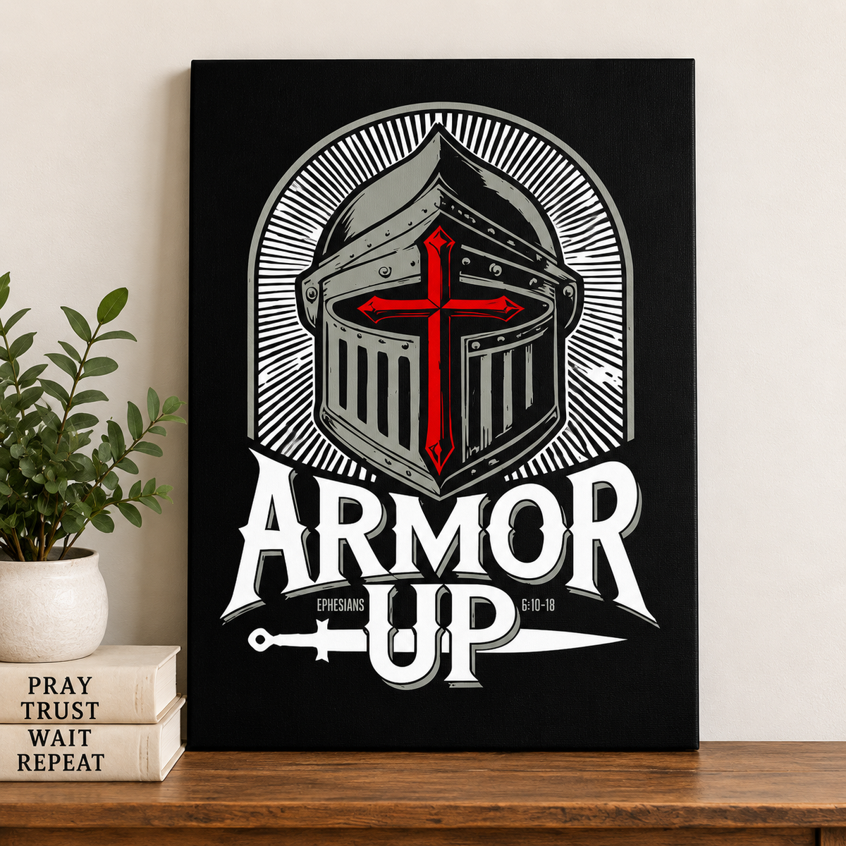 A black canvas wall art print featuring a stylized medieval knight helmet with a red cross, the words 'ARMOR UP,' and a small 'EPHESPHAN' text, displayed leaning on a wooden shelf next to a potted plant and stacked books.