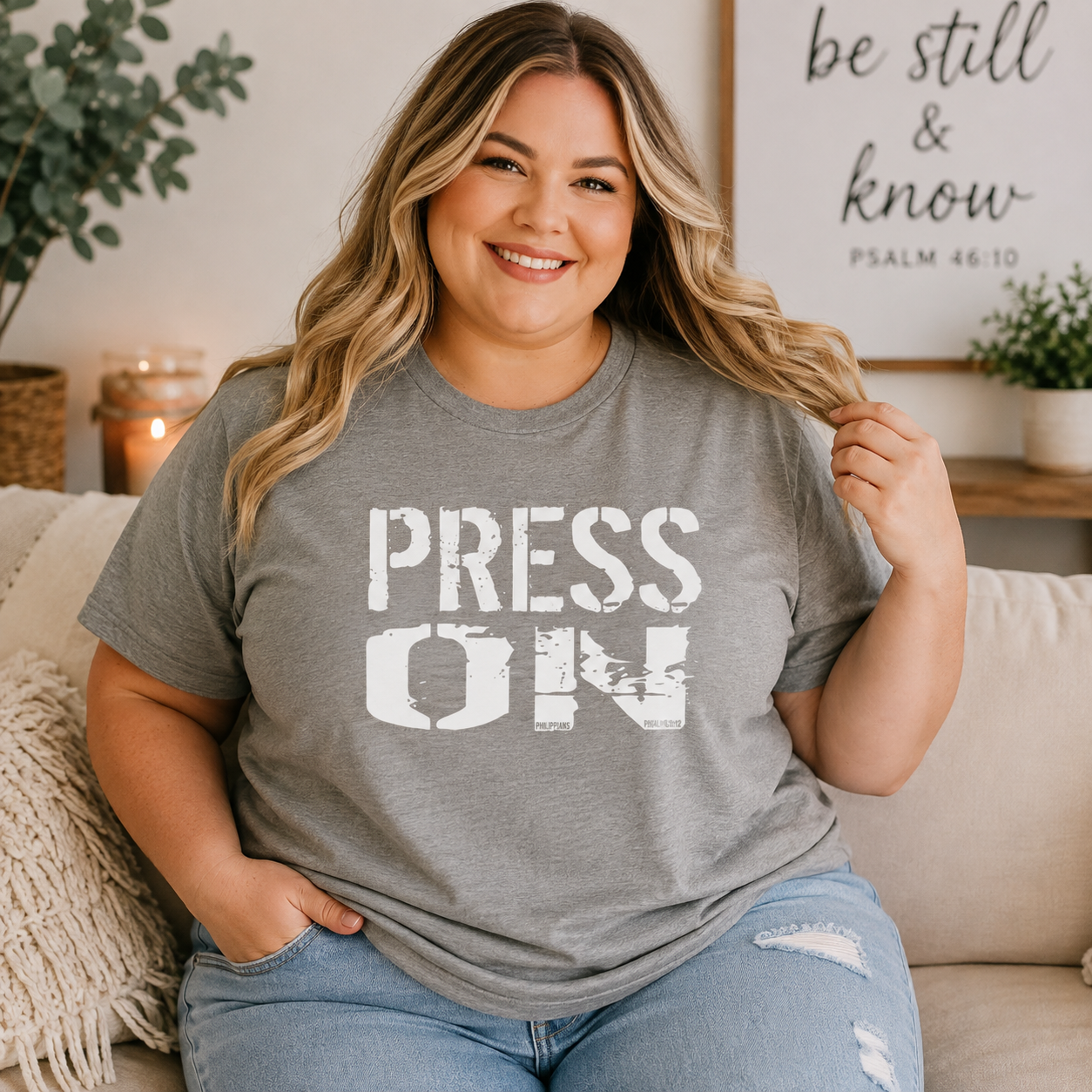 A woman sitting on a beige couch wearing a heather gray t-shirt with the words 'PRESS ON' printed in distressed white block letters. She is wearing light blue ripped jeans and smiling, with a cozy home background featuring a framed sign that reads 'be still & know PSALM 46:10'.
