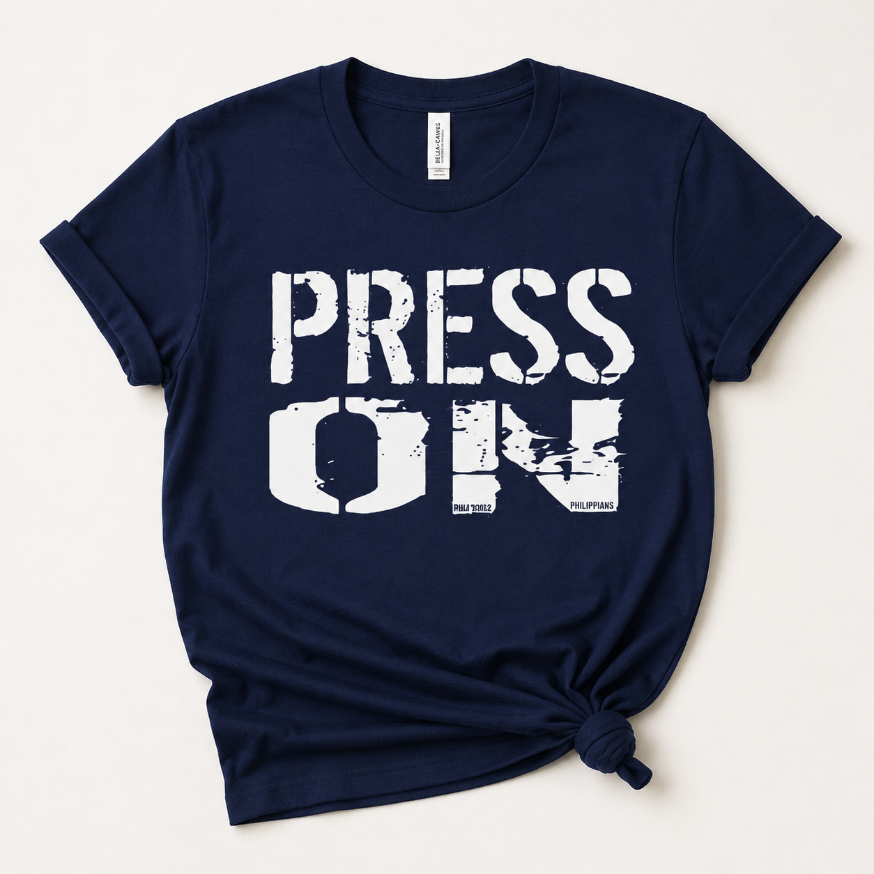 A navy blue short-sleeve t-shirt with 'PRESS ON' printed in distressed white block letters across the front, styled with a knot at the bottom hem on a light beige background.