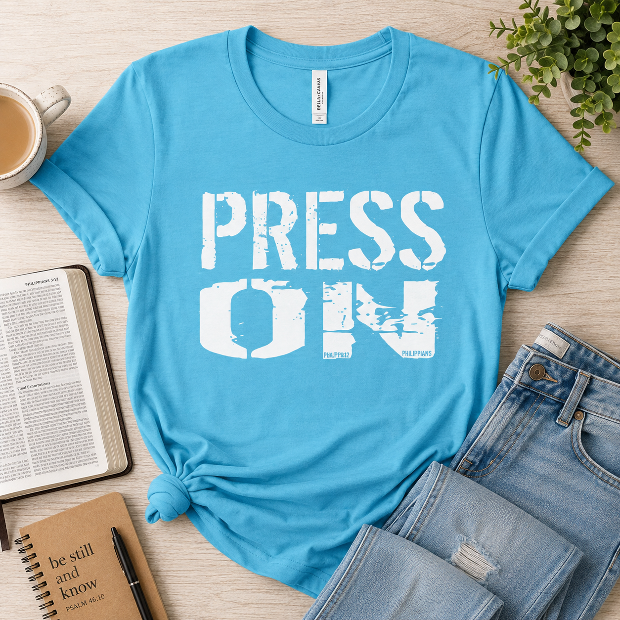 A bright blue graphic T-shirt laid flat on a light wood surface with a coffee cup, book, notebook, and jeans around it. The shirt has large distressed white text reading 'PRESS ON' across the front.