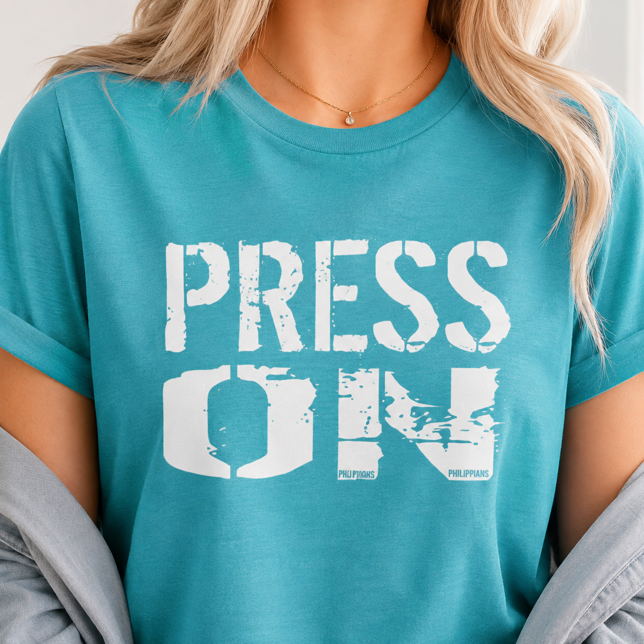 A woman wearing a teal t-shirt with a distressed white graphic that reads 'PRESS ON' with 'PHILIPPINES' printed below the letters. She has blonde hair and is wearing a gold necklace.