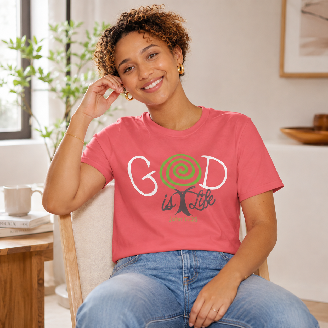 A woman sitting in a chair wearing a coral pink graphic t-shirt with 'GOD is Life' printed on the front, paired with blue jeans in a bright, modern living room setting.