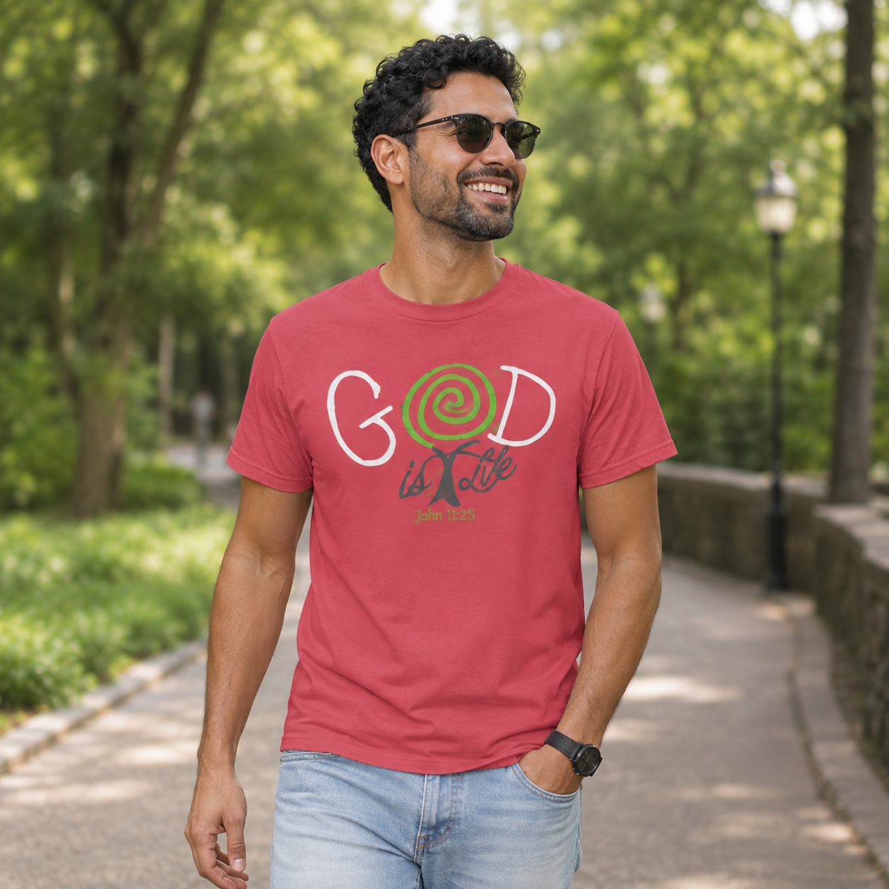 A man wearing a coral red graphic t-shirt with 'GOD is Love' printed on the front, walking on a paved path in a park with green trees in the background.