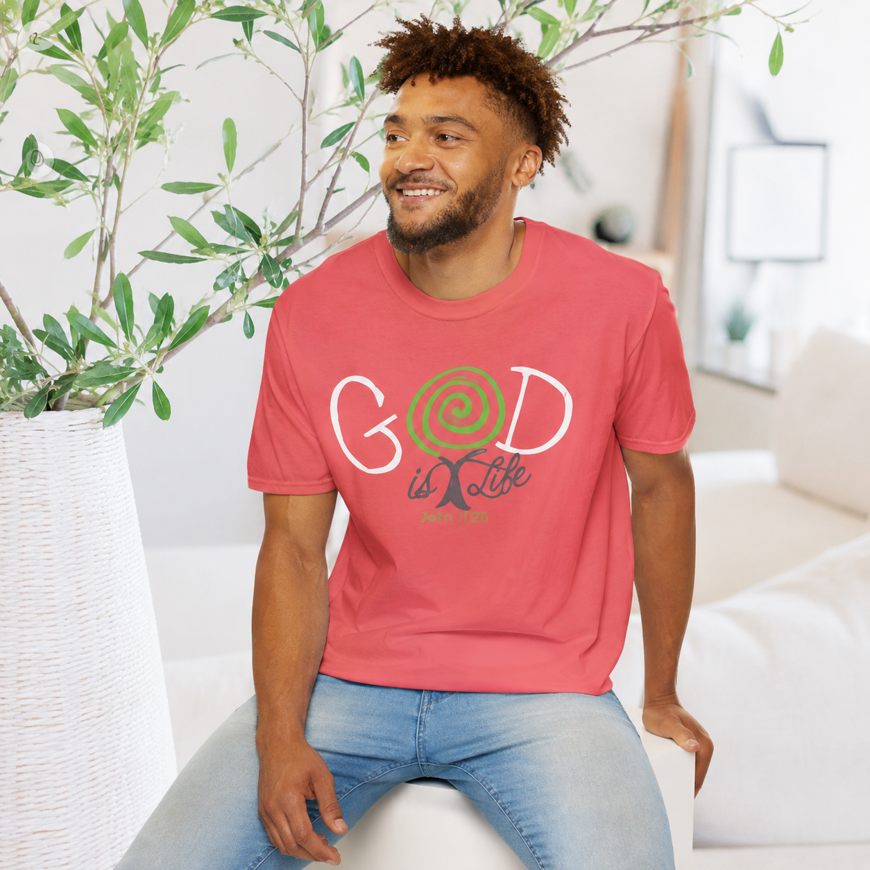 A man wearing a coral pink t-shirt with a 'God is Life' graphic, sitting in a bright, modern living room.