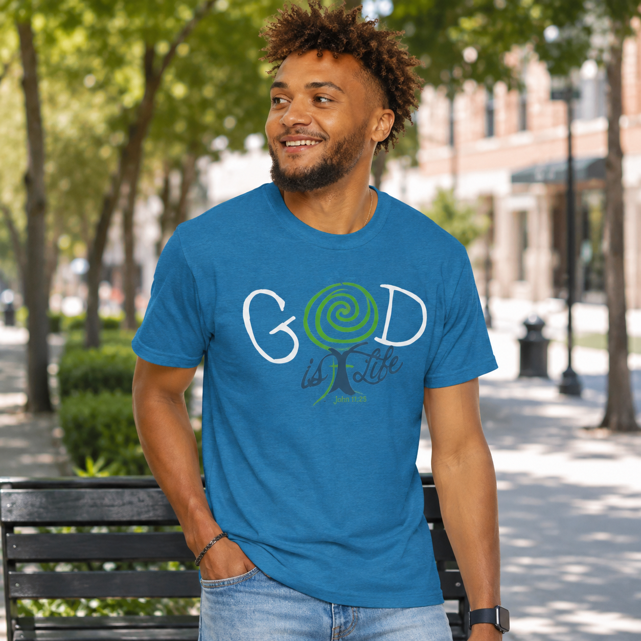 A man wearing a blue graphic t-shirt with 'GOD is Love' printed on the front, standing on a park bench with trees and a city street in the background.
