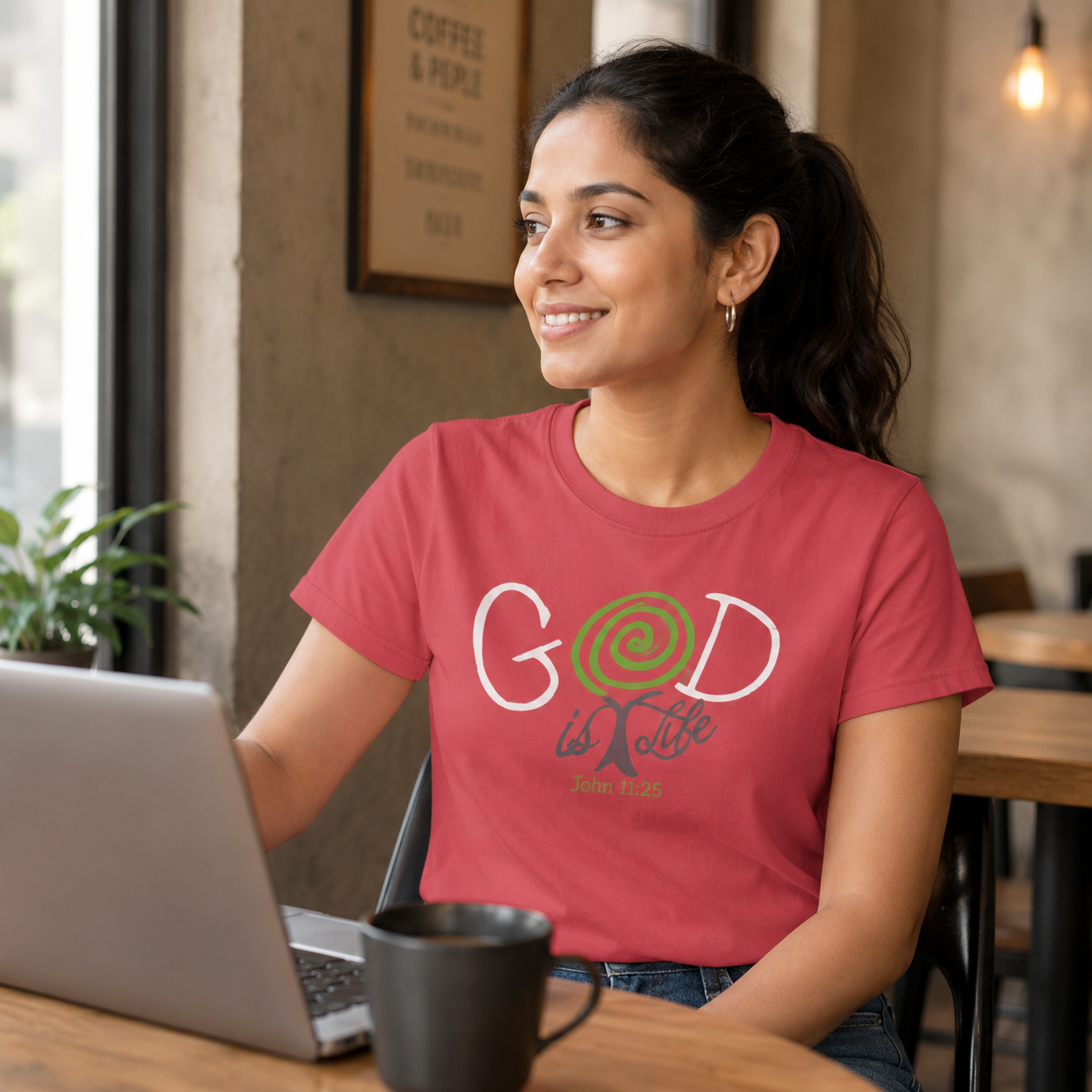 A woman sitting at a cafe table wearing a coral pink t-shirt with a graphic that reads 'God is Life' in white and green. She is smiling and looking to the side while using a laptop.