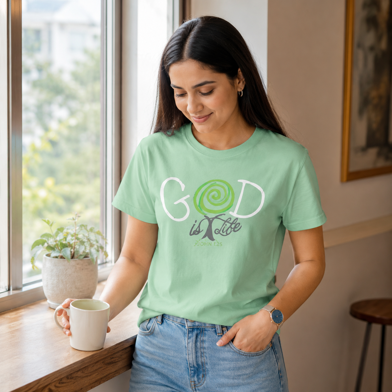 A woman wearing a light green graphic t-shirt with 'GOD is life' printed on the front, standing by a window and holding a mug.