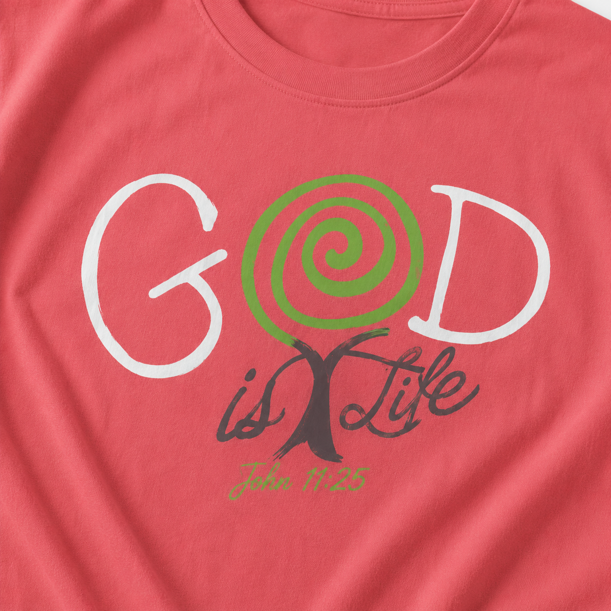 A close-up of a coral pink t-shirt featuring a graphic print that reads 'GOD is Life' with 'John 1:25' underneath. The letter 'O' is replaced by a green spiral, and the letter 'I' is replaced by a dark brown tree trunk.