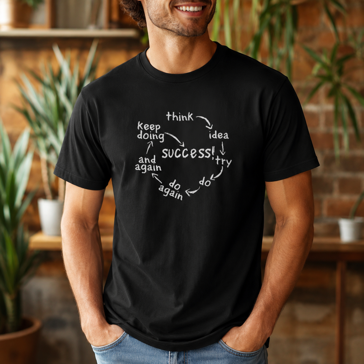 A man wearing a black crewneck t-shirt with a white circular graphic that reads 'think keep doing and again success! try do do again'. The background is a warm, blurred indoor setting with brick walls and potted plants.
