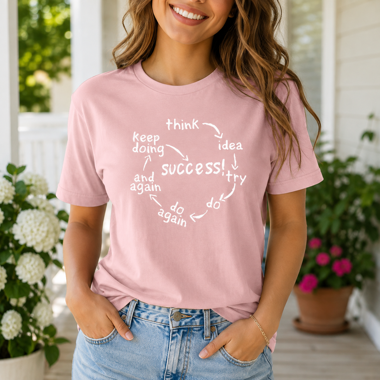 A woman wearing a light pink graphic t-shirt with a white circular motivational quote about success, paired with light blue jeans, standing on a porch with potted plants in the background.