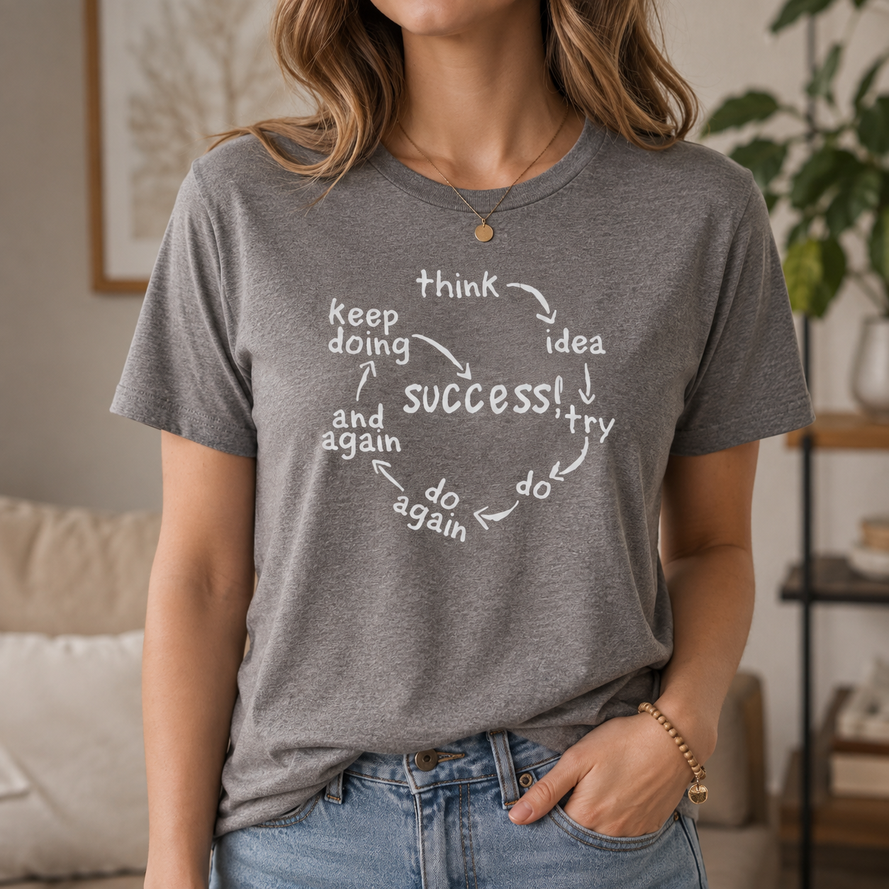 A woman wearing a heather gray graphic t-shirt with a white circular motivational quote that reads 'think idea try do again success! and again'. The background shows a softly blurred living room interior with a couch and a plant.