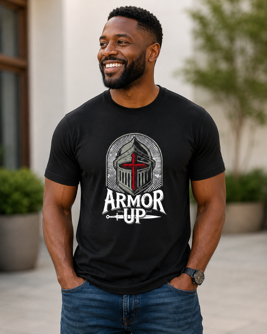 A smiling man wearing a black graphic t-shirt with a knight helmet and the words 'ARMOR UP' in white, gray, and red. He is wearing blue jeans and standing outdoors in front of a blurred building background.